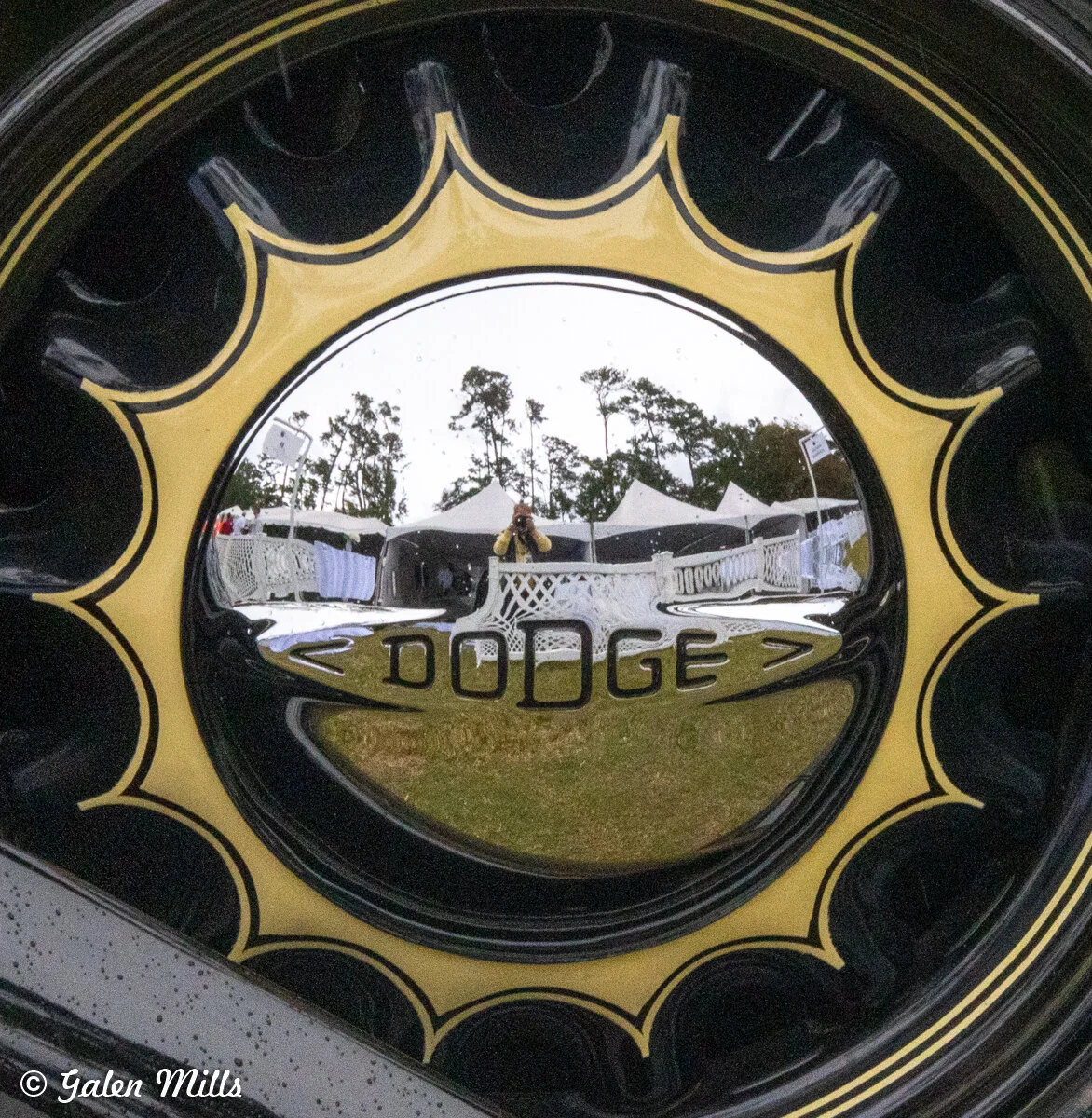 Close-up of a vintage Dodge hubcap with reflective surface showing photographer's reflection and outdoor setting.