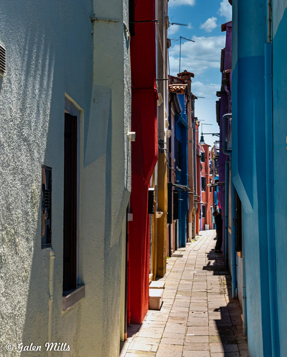 Narrow alleyway with colorful painted buildings, featuring blue, red, and yellow walls, under a blue sky with clouds.