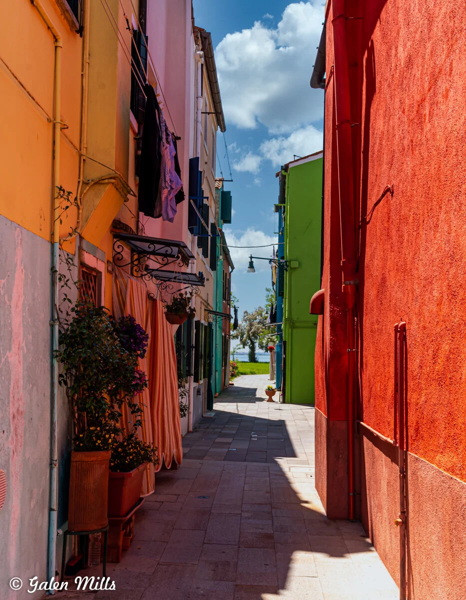 Colorful alleyway with brightly painted buildings in shades of yellow, green, and red. A narrow path leads to trees and distant water, under a partly cloudy sky. Hanging plants and laundry adorn the walls.