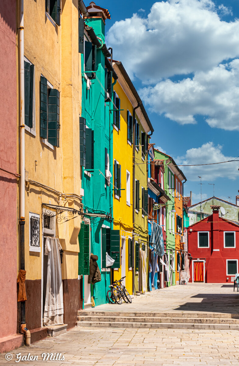 Colorful houses with green shutters on a sunny street with bicycles and laundry hanging, under a blue sky with clouds.