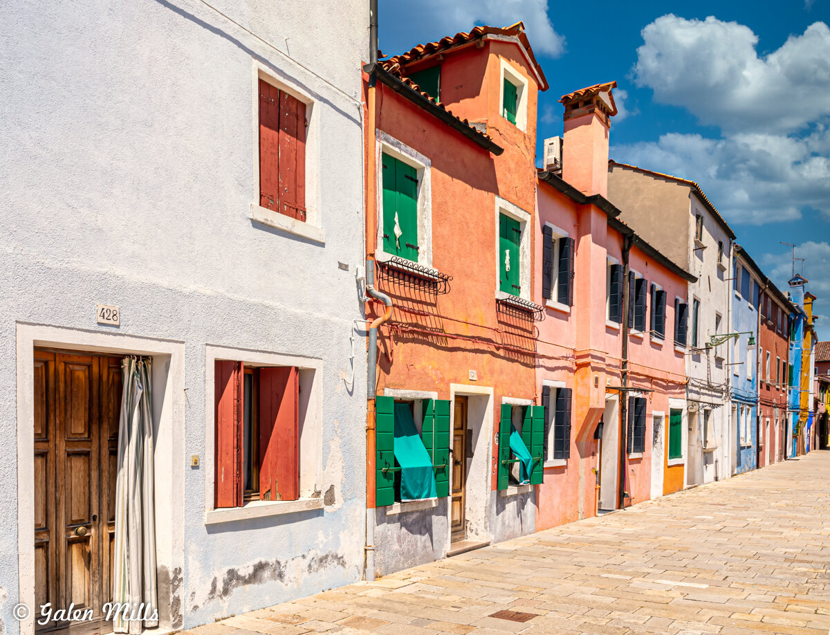 Colorful houses along a sunny street in Burano, Italy, featuring pastel walls and vibrant shutters.