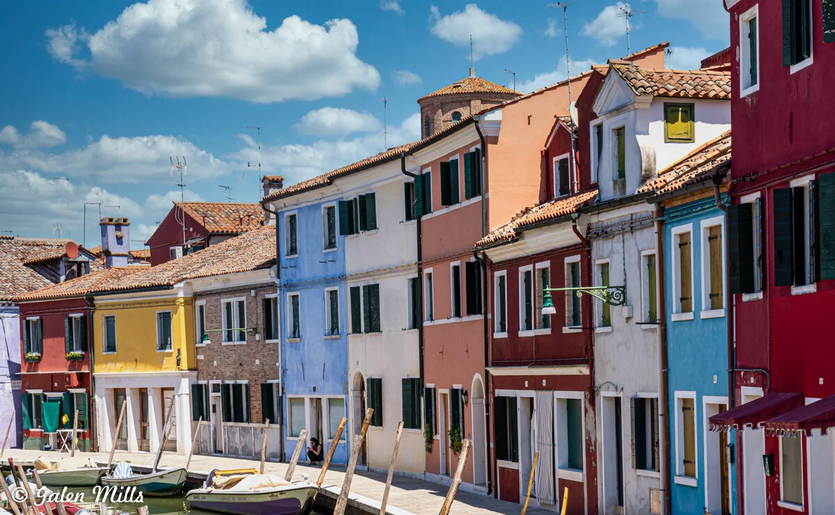 Colorful houses along a canal in Burano, Italy, under a blue sky with clouds.