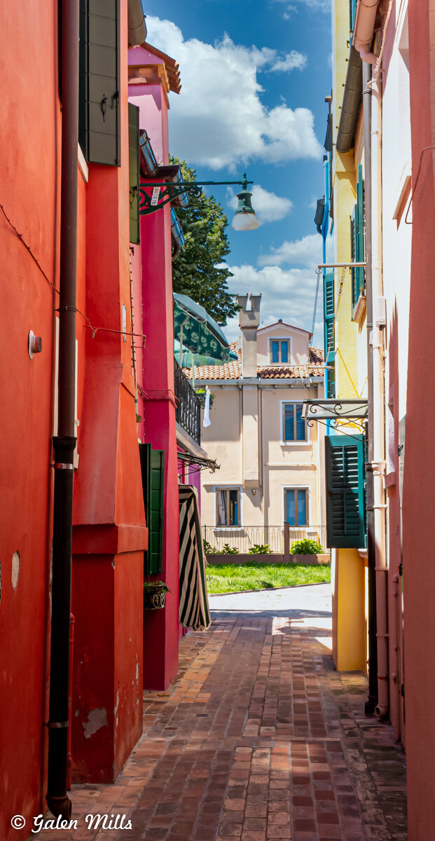 Narrow alleyway with colorful buildings, red and pink walls, shutters, brick walkway, and view of a pastel building with blue shutters under a blue sky.