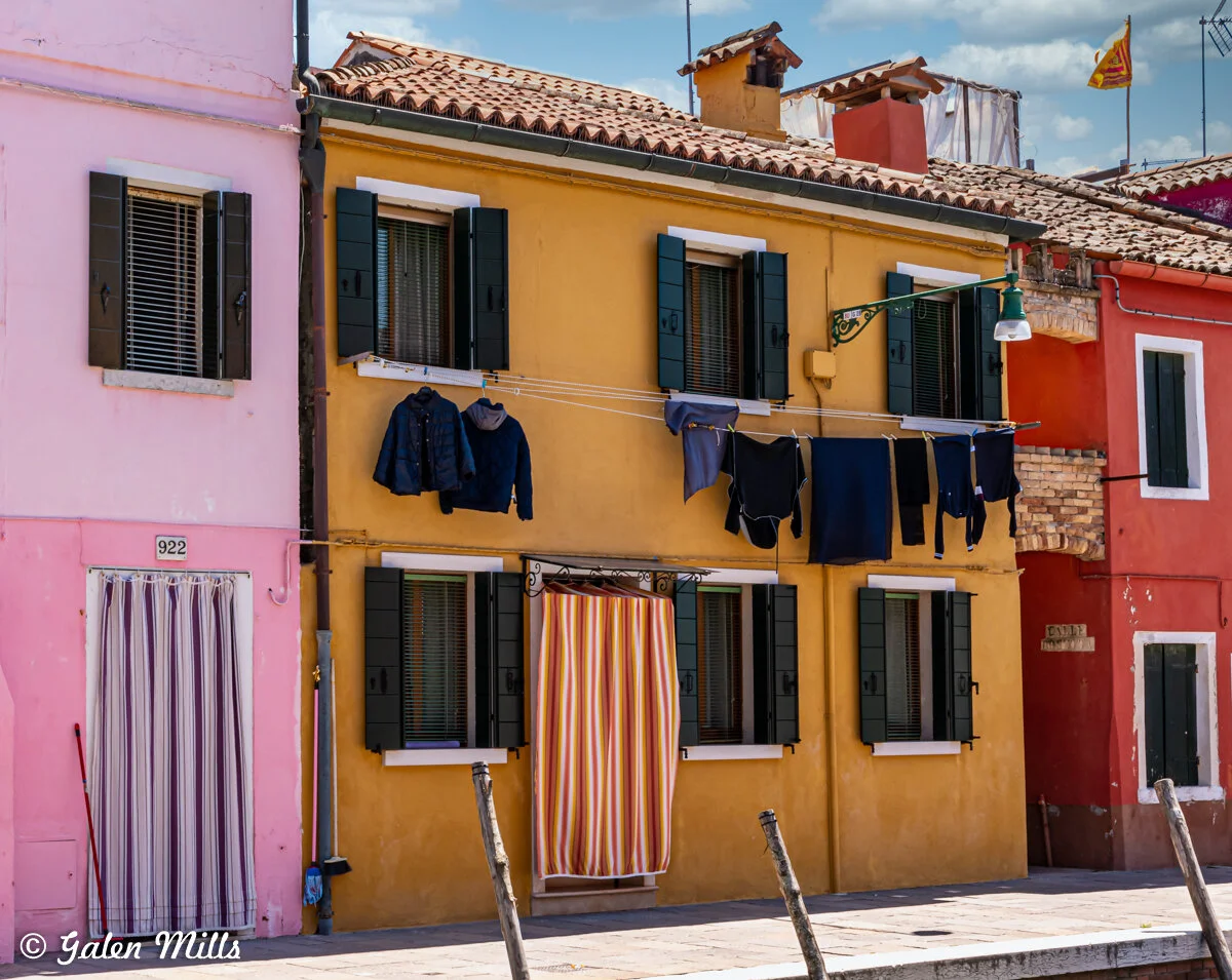 Colorful houses with laundry hanging on lines in Burano, Italy, featuring pink and yellow facades, green shutters, and striped curtains.