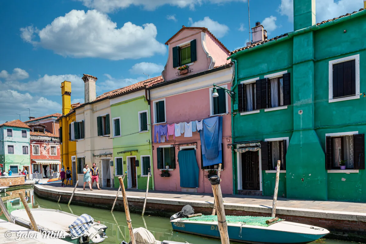 Colorful buildings along a canal in Burano, Italy, with laundry hanging outside and boats moored nearby.