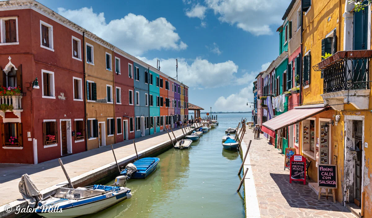 Colorful canal in Burano, Italy, with boats moored along the water and bright buildings on both sides under a blue sky.