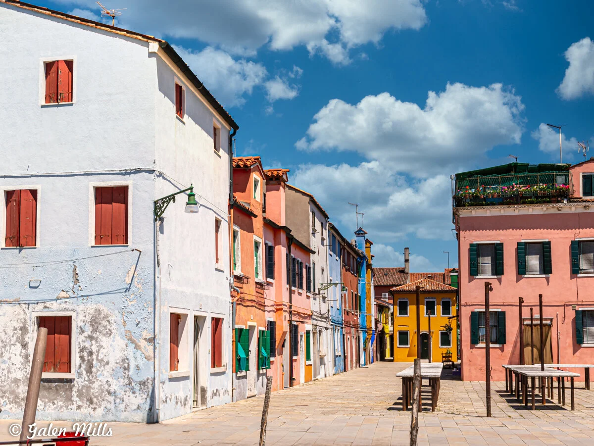 Colorful buildings with closed shutters lining a cobblestone street under a blue sky with clouds.