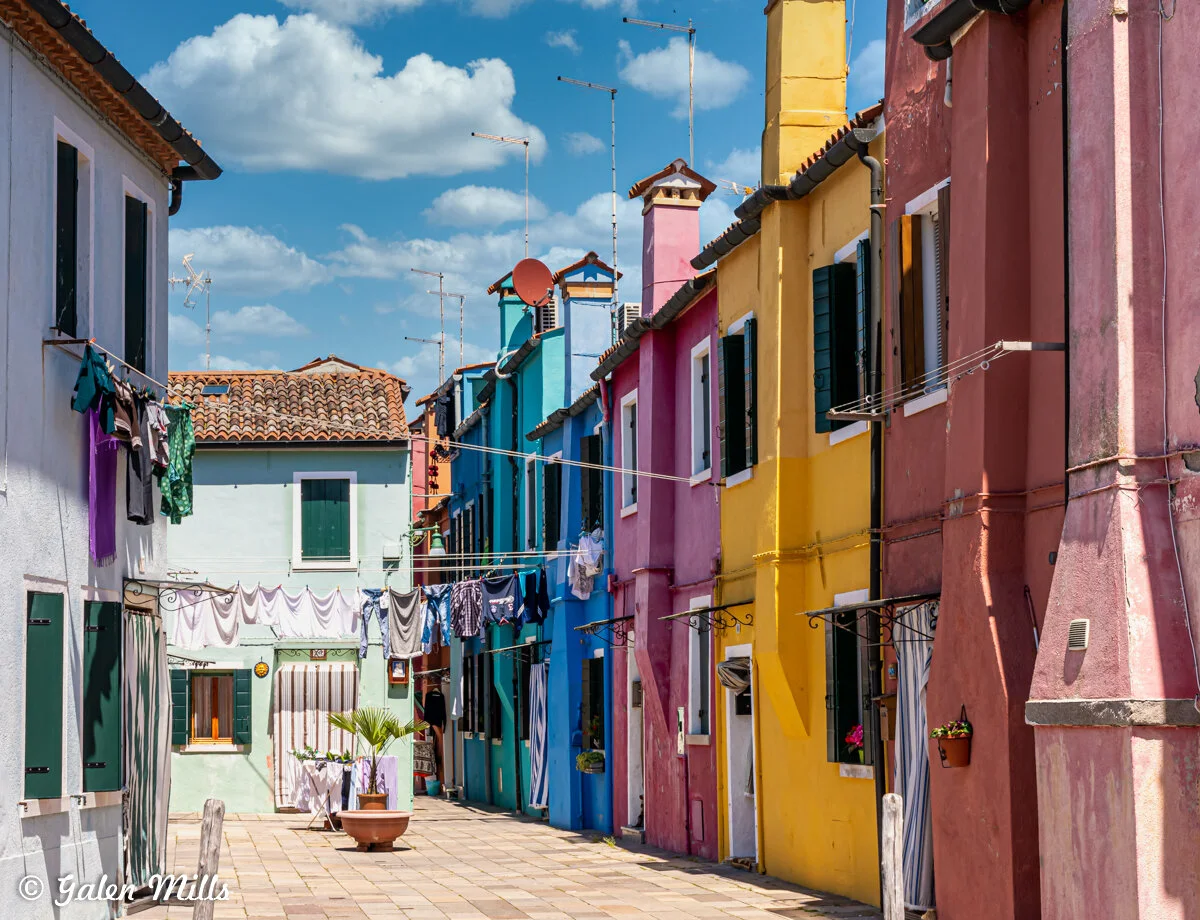 Colorful houses along a narrow cobblestone street in Burano, Italy, with laundry hanging on clotheslines between the buildings.