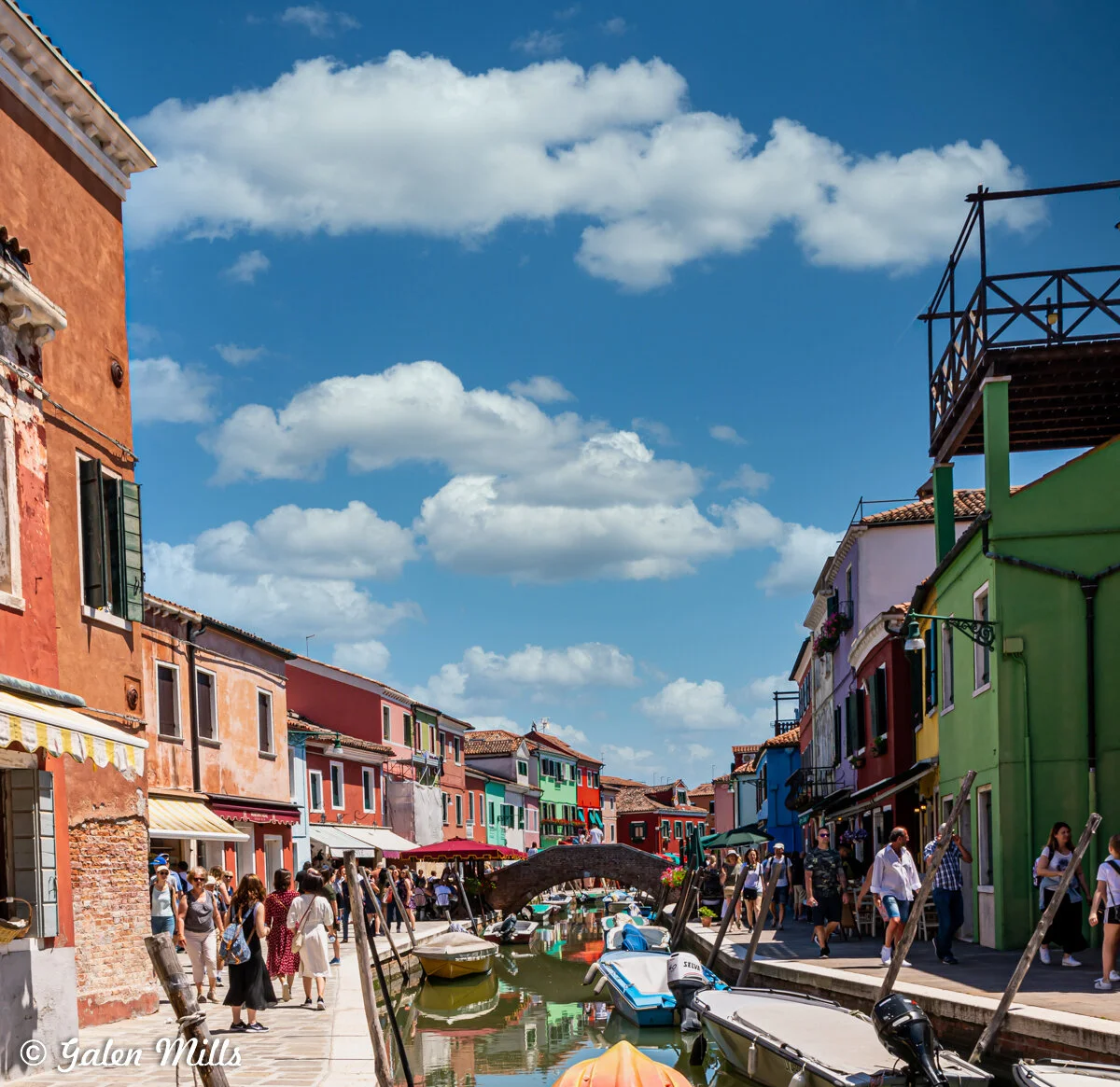 Colorful buildings line a canal in Burano, Italy, with boats moored along the waterway. People walk along the street and a small arched bridge spans the canal. The sky is clear with some clouds.