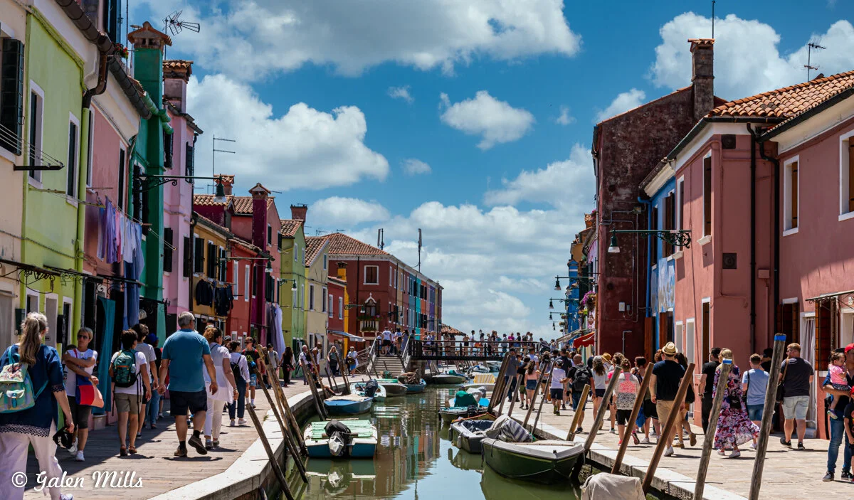 Colorful houses and canal in Burano, Italy with people walking along the promenade and boats docked on the water.