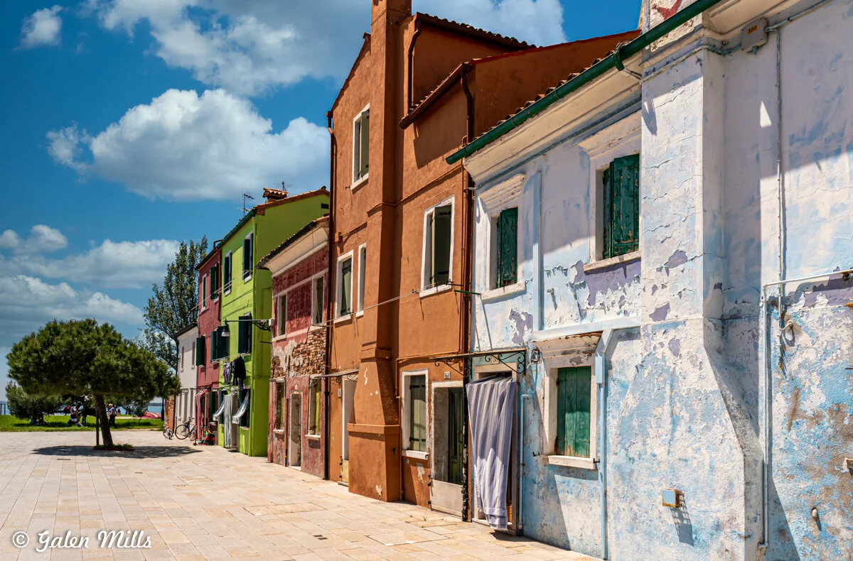 Colorful buildings with peeling paint in Burano, Italy, under a sunny sky with fluffy clouds.