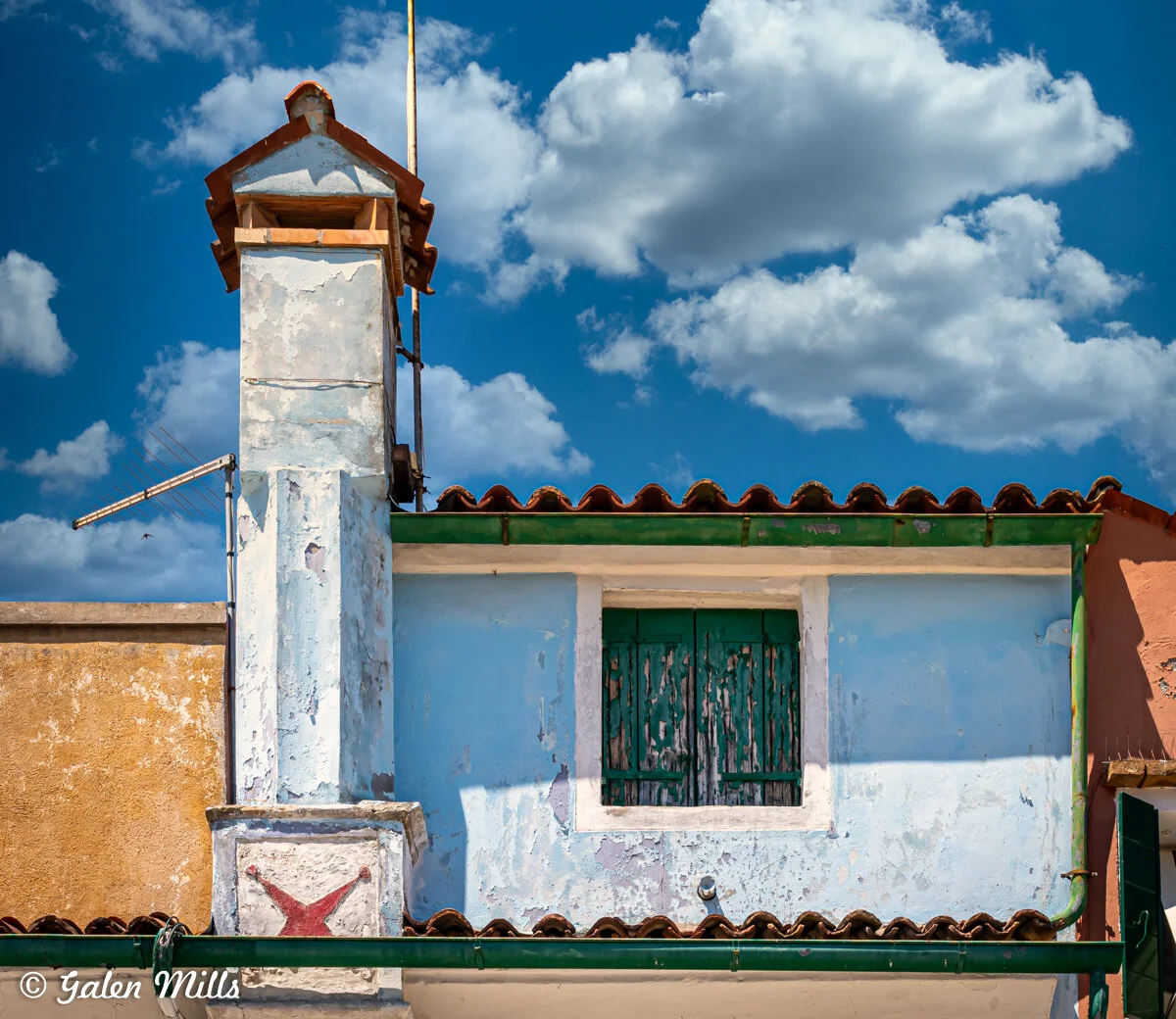 Old building with a weathered blue facade, a chimney, and a green shuttered window under a cloudy sky.