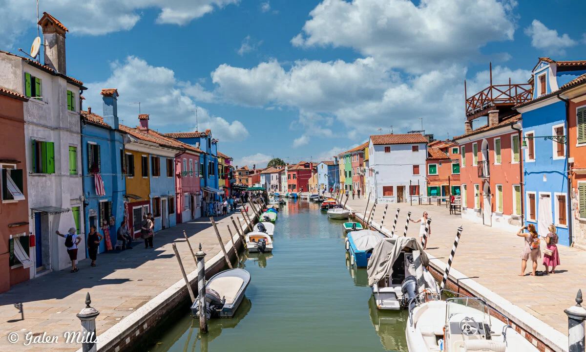 Colorful houses along a canal in Burano, Italy with boats docked and people walking on a sunny day.