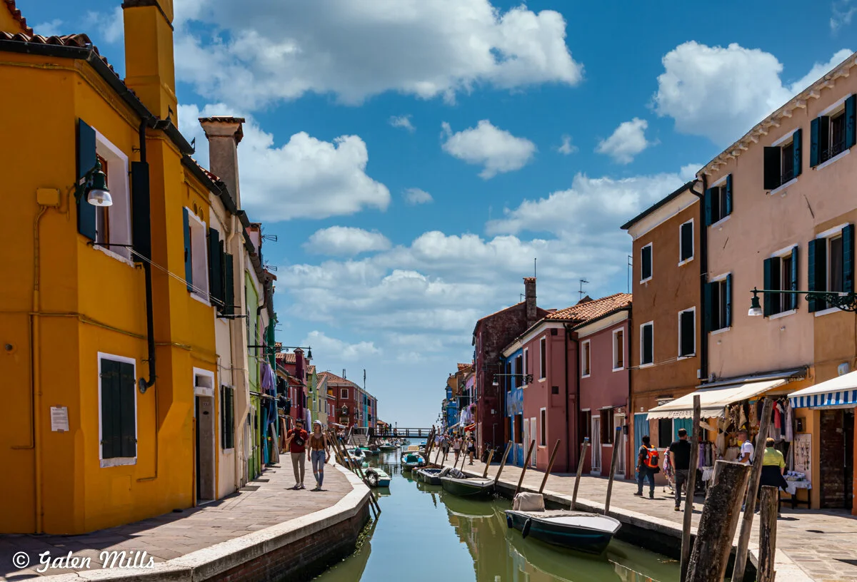 Colorful houses line a canal with boats in Burano, Italy, under a bright blue sky.