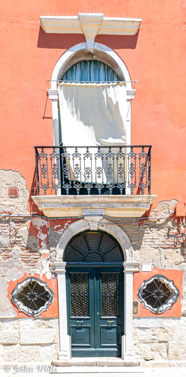 A building facade featuring an arched entrance door with decorative ironwork, flanked by two grille-covered windows. Above is a small balcony with ornate black iron railings and an arched window covered by a curtain. The facade shows signs of aging w