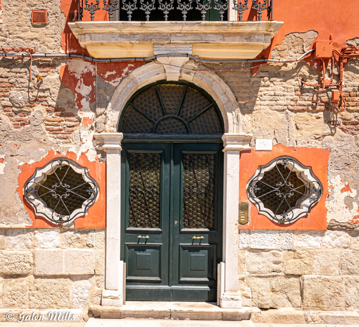 A historic building facade featuring a large arched, dark green double door with ornate glass, surrounded by a stone and brick exterior. The building has two uniquely shaped, decorative windows on either side of the door, each with a patterned metal 