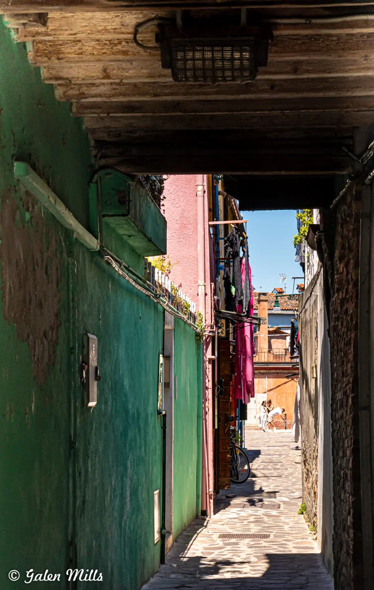 Narrow alley with colorful buildings, clothes hanging, and a bicycle; light filtering from above.