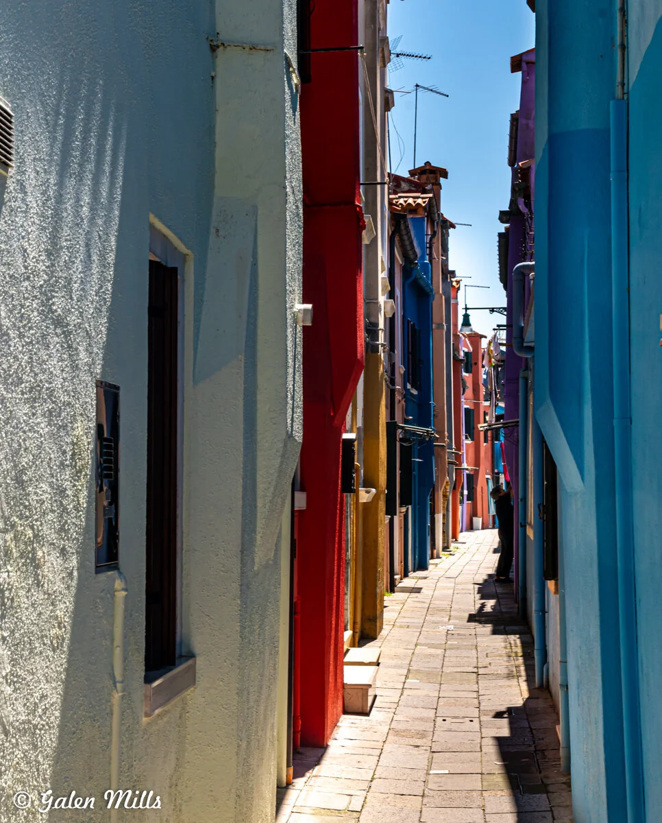 Narrow alley with colorful buildings, including blue, red, and yellow walls, casting shadows on the cobblestone path below. Bright, sunny day with clear sky and some antennas visible.