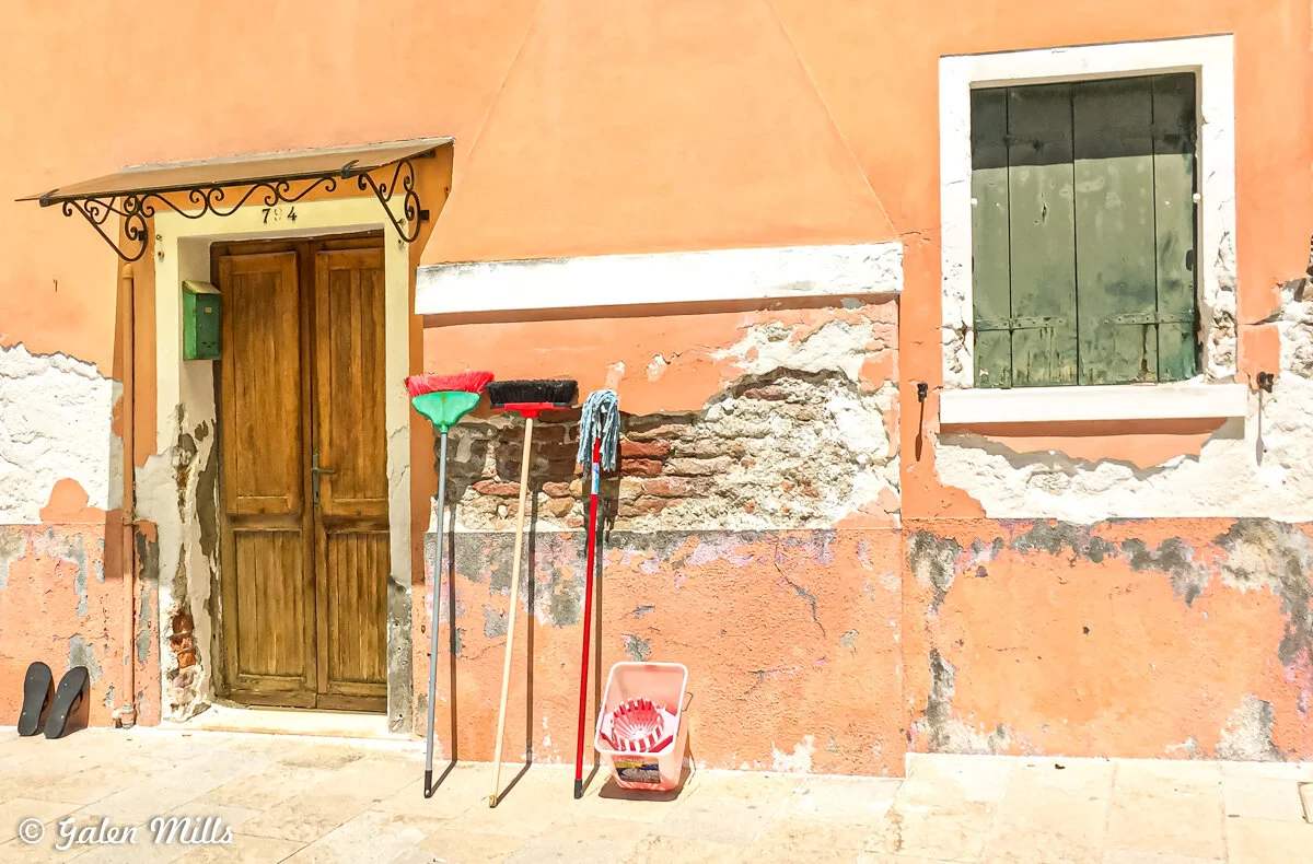 Weathered orange wall with a wooden door, green mailbox, two brooms, mop, bucket, and shuttered window.