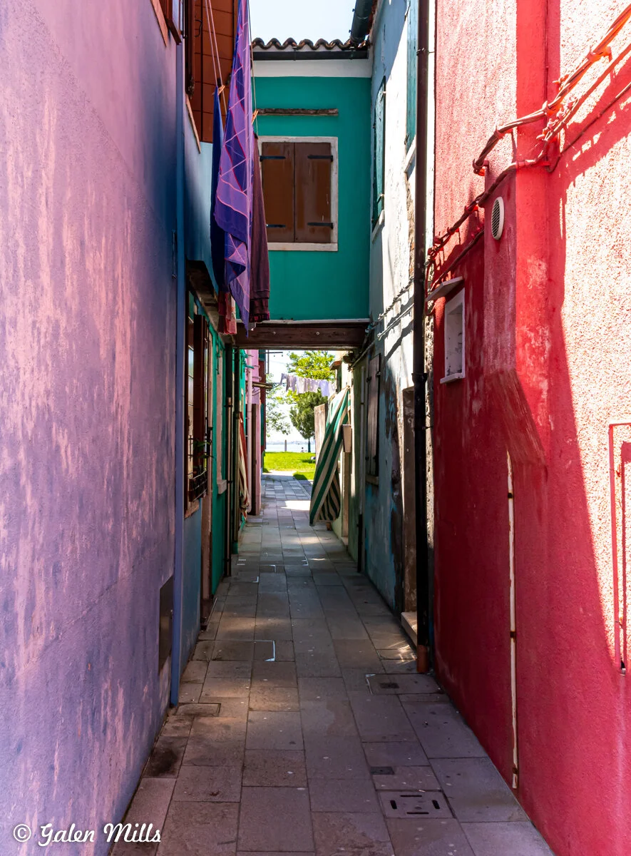 Narrow alleyway with colorful buildings and laundry hanging, leading to a view of greenery in the distance.