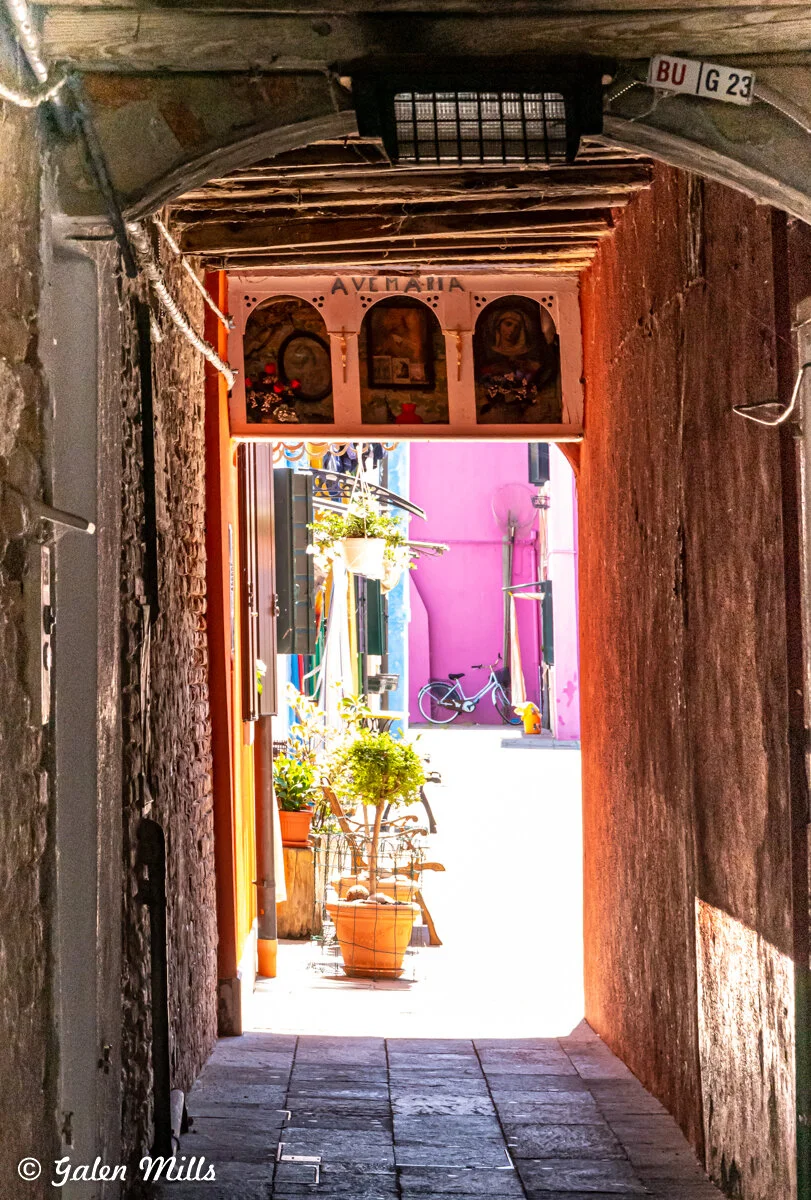 A narrow stone corridor leading to a colorful courtyard with potted plants and a bicycle. The entrance features religious icons and the words "Ave Maria." Sunlight illuminates the vivid pink and blue buildings outside.