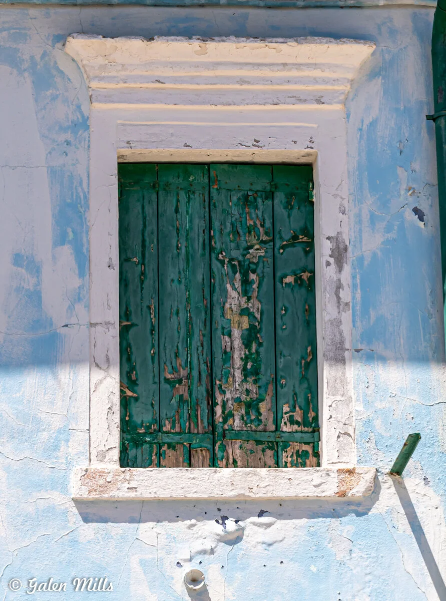 Weathered green wooden shutters on a faded blue stucco wall with peeling paint and a small pipe below.