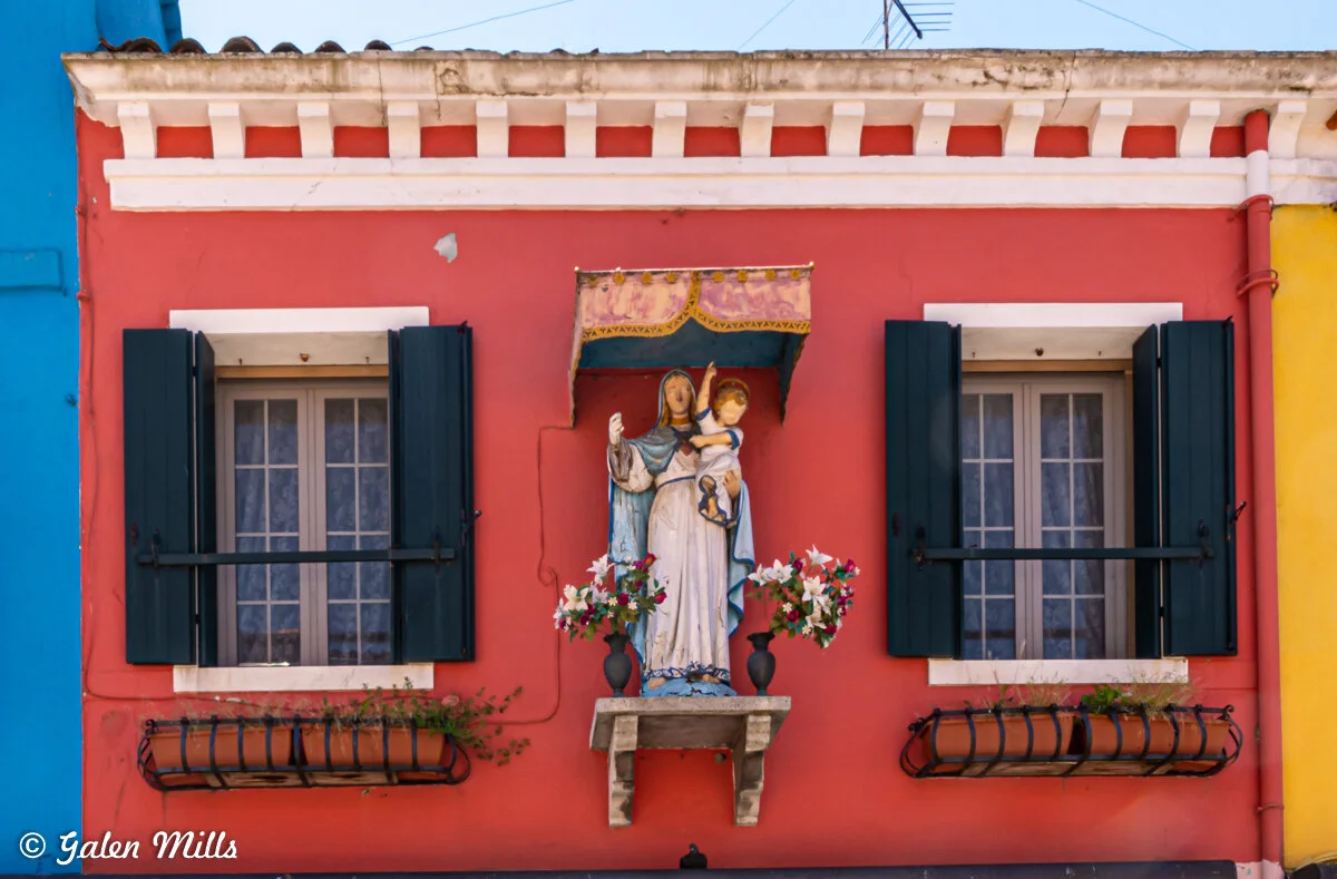Colorful building facade with a religious statue, flowers, and windows in Burano, Italy.