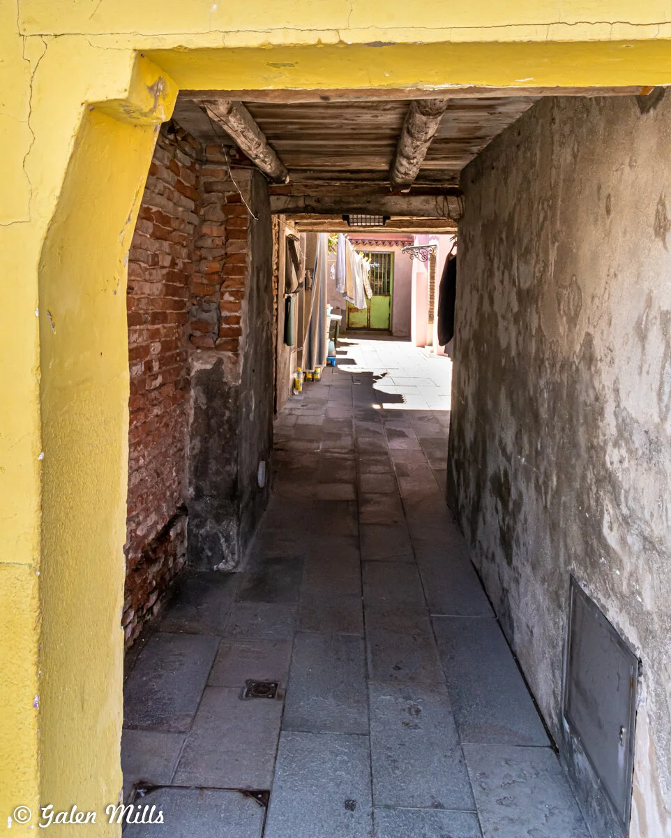 Narrow alleyway with yellow walls, stone floor, and wooden ceiling leading to a bright courtyard featuring laundry hanging outside.