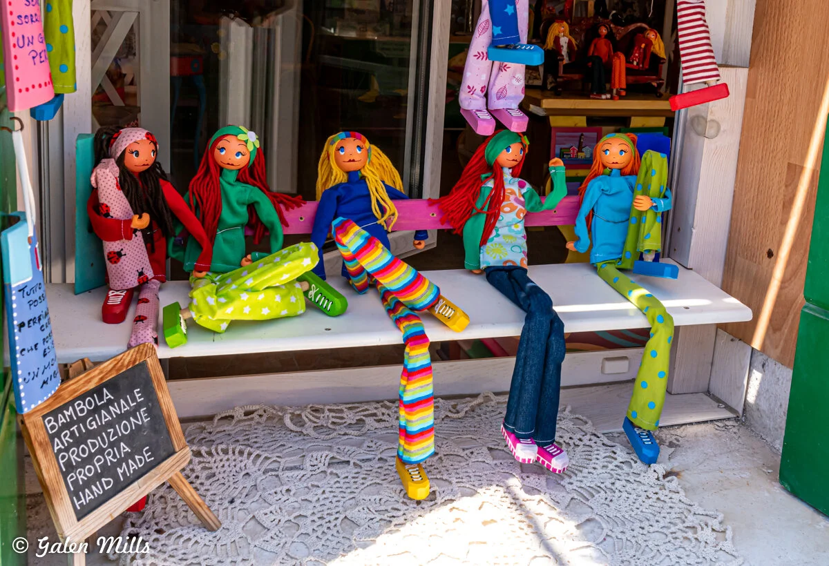Handmade cloth dolls sitting on a display bench in a shop, featuring colorful and patterned fabrics, with a wooden sign indicating handmade artisanal production.