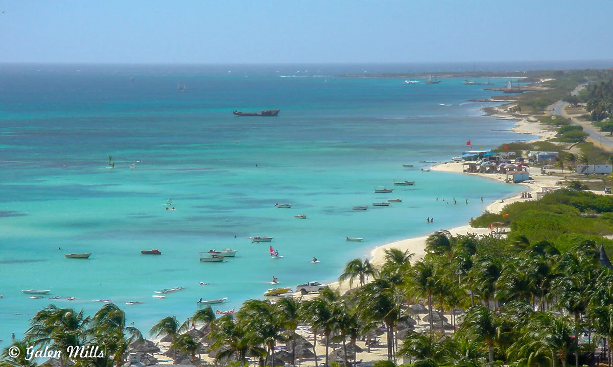 A tropical beach with turquoise water, boats, and palm trees along the shore. People are swimming and relaxing on the beach. The coastline stretches into the distance, with a road and buildings visible.