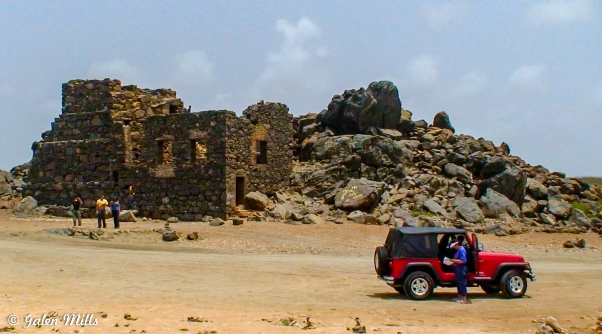 Old stone building and rocks with people nearby, next to a red Jeep in a desert landscape.