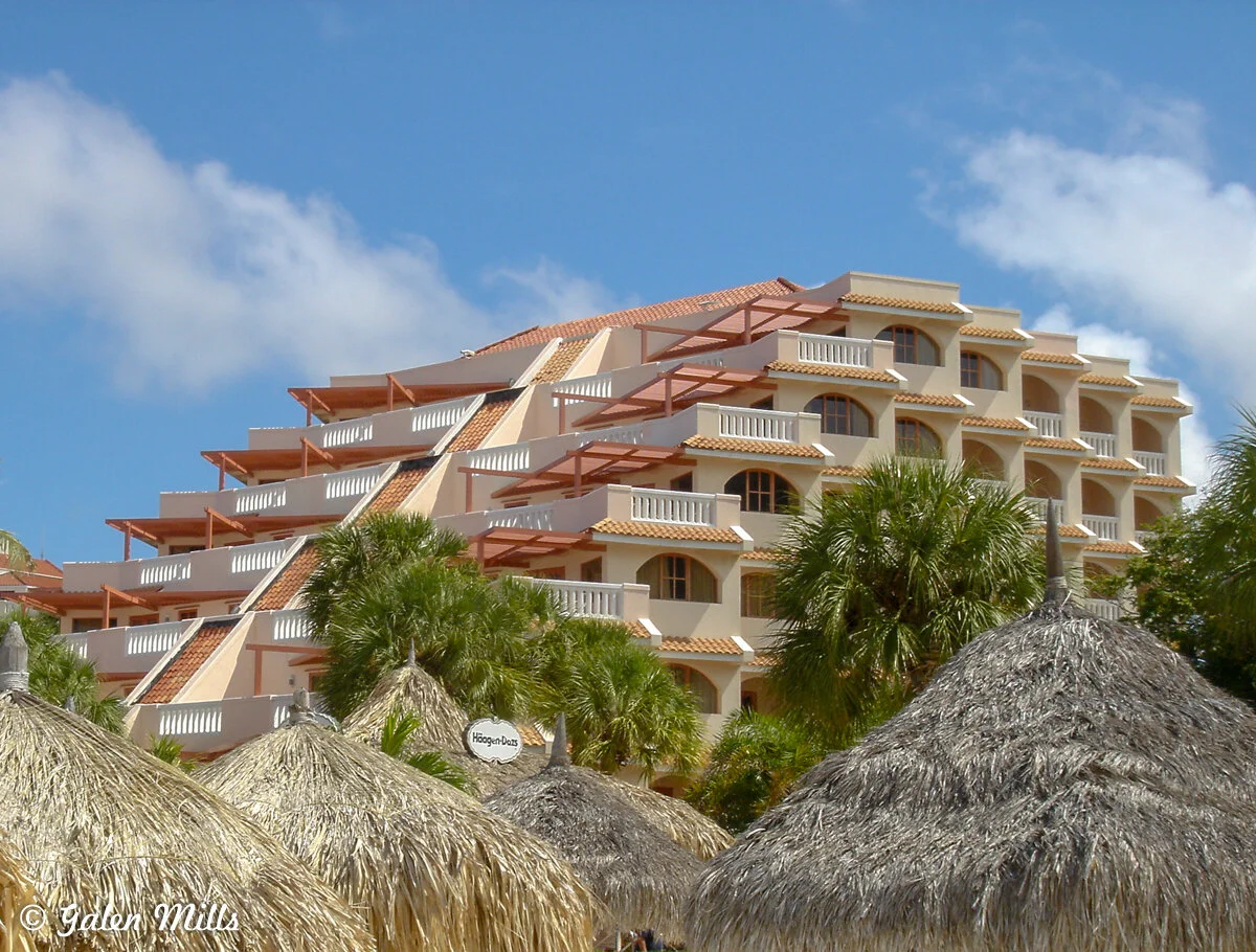 Hotel building with palm trees and straw thatched huts against blue sky