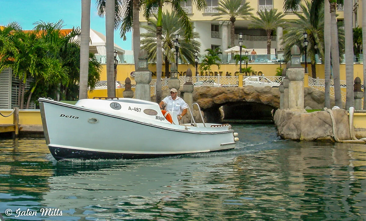 Small boat with a captain docking near a resort, surrounded by palm trees and a stone bridge, sunny day.