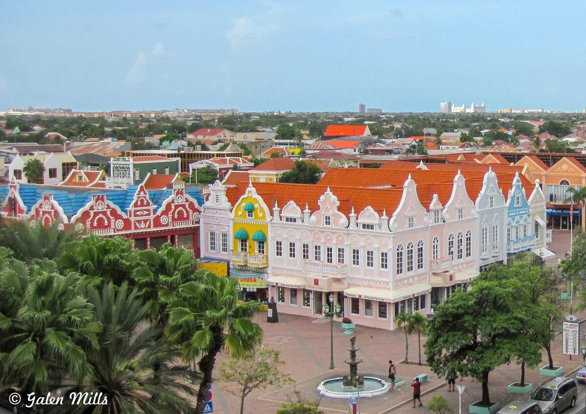 Colorful Dutch colonial buildings with vibrant red, blue, yellow, and pink facades in Oranjestad, Aruba, with palm trees and a small fountain in the foreground.