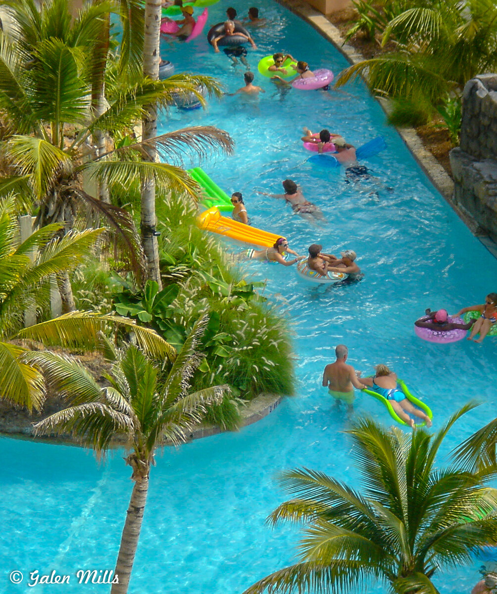 People enjoying a lazy river at a tropical resort with palm trees and various inflatable pool floats.