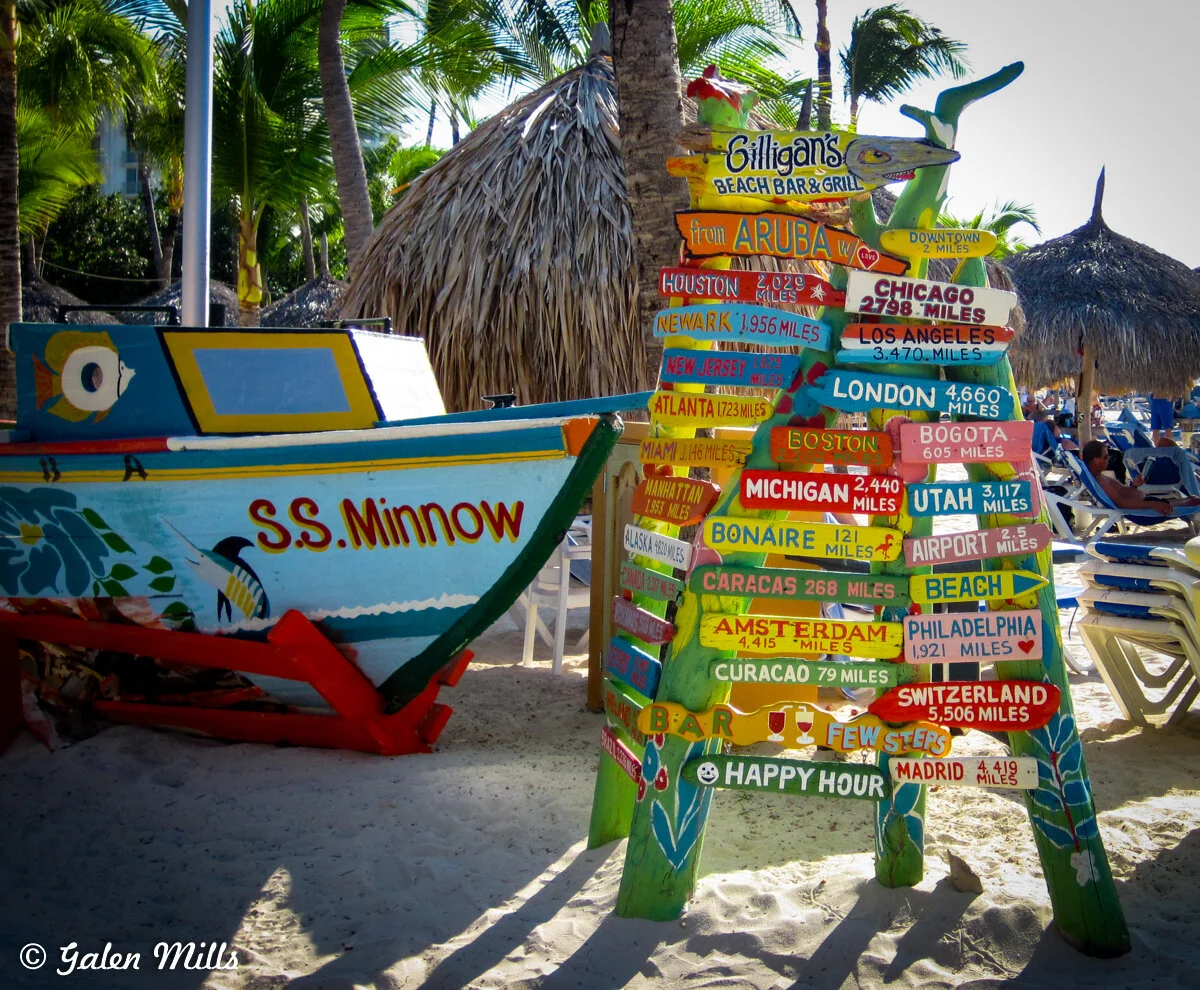 Colorful directional signpost on a beach with distances to various cities, next to a small boat labeled 'S.S. Minnow.'
