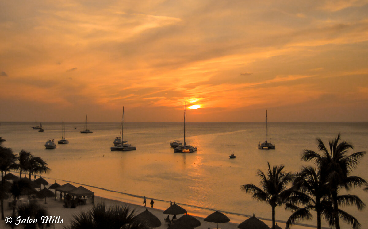 Sunset over tropical beach with sailboats on the water, palm trees, and straw umbrellas.