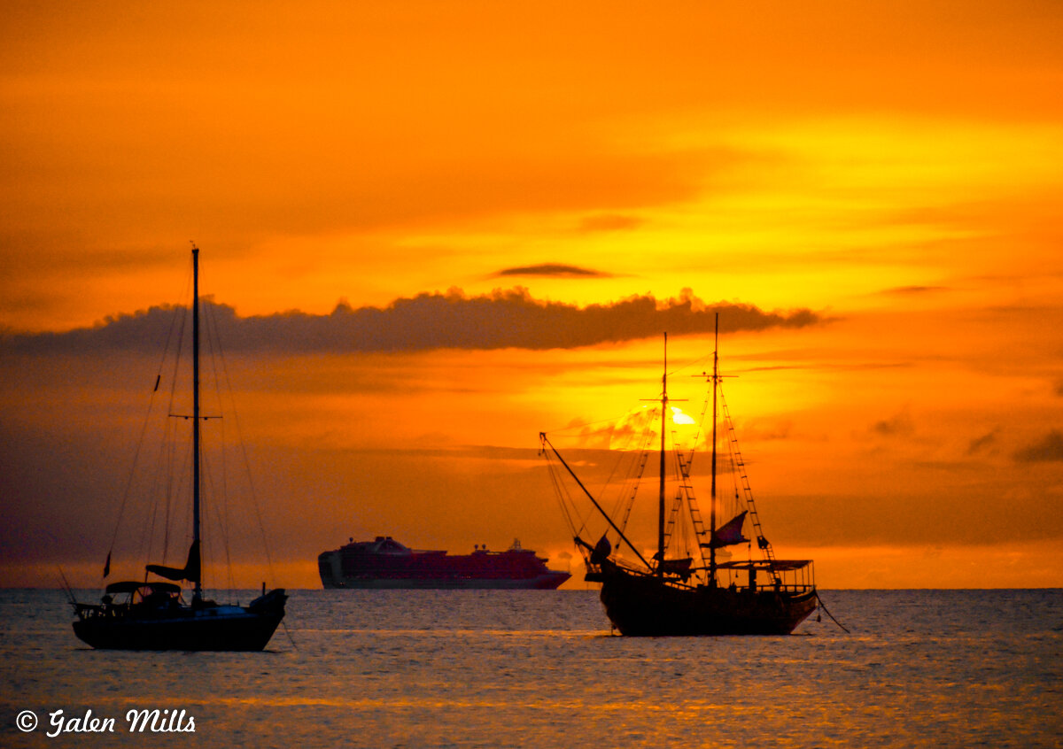 Silhouettes of two sailboats and a cruise ship on the ocean at sunset, with an orange sky.
