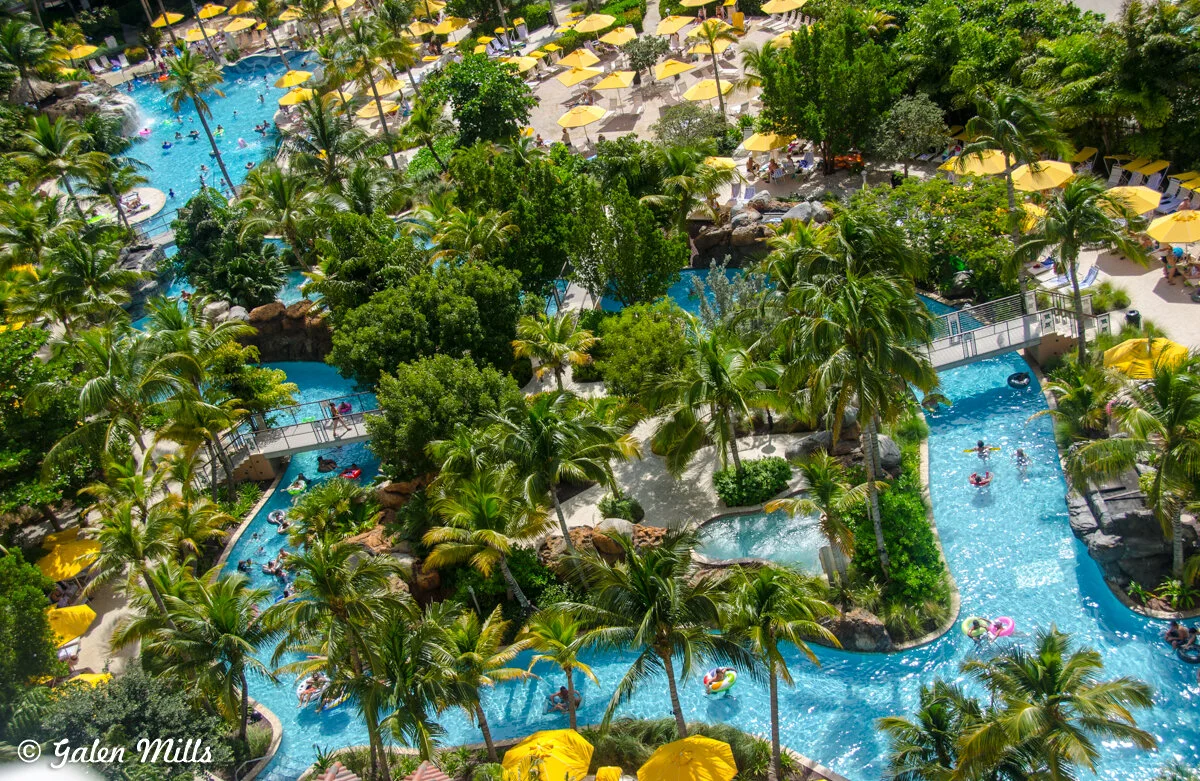 Aerial view of a tropical resort featuring a winding blue pool with people swimming. Surrounded by lush green foliage, palm trees, and yellow sun umbrellas on a sunny day.