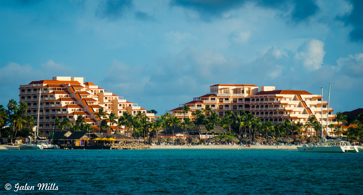 Resort buildings with red roofs, surrounded by palm trees, next to a beach and ocean with blue water and moored boats.