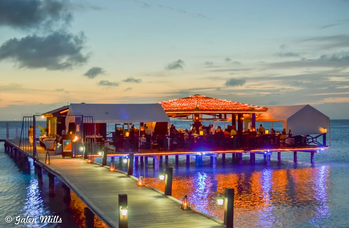 Waterfront restaurant at sunset with string lights and dining area on a pier over water.