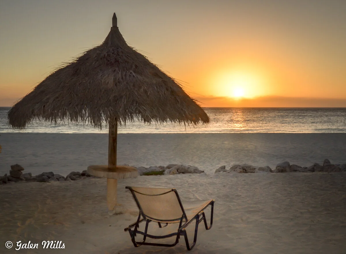 Beach at sunset with a thatched umbrella and lounge chair on the sand.