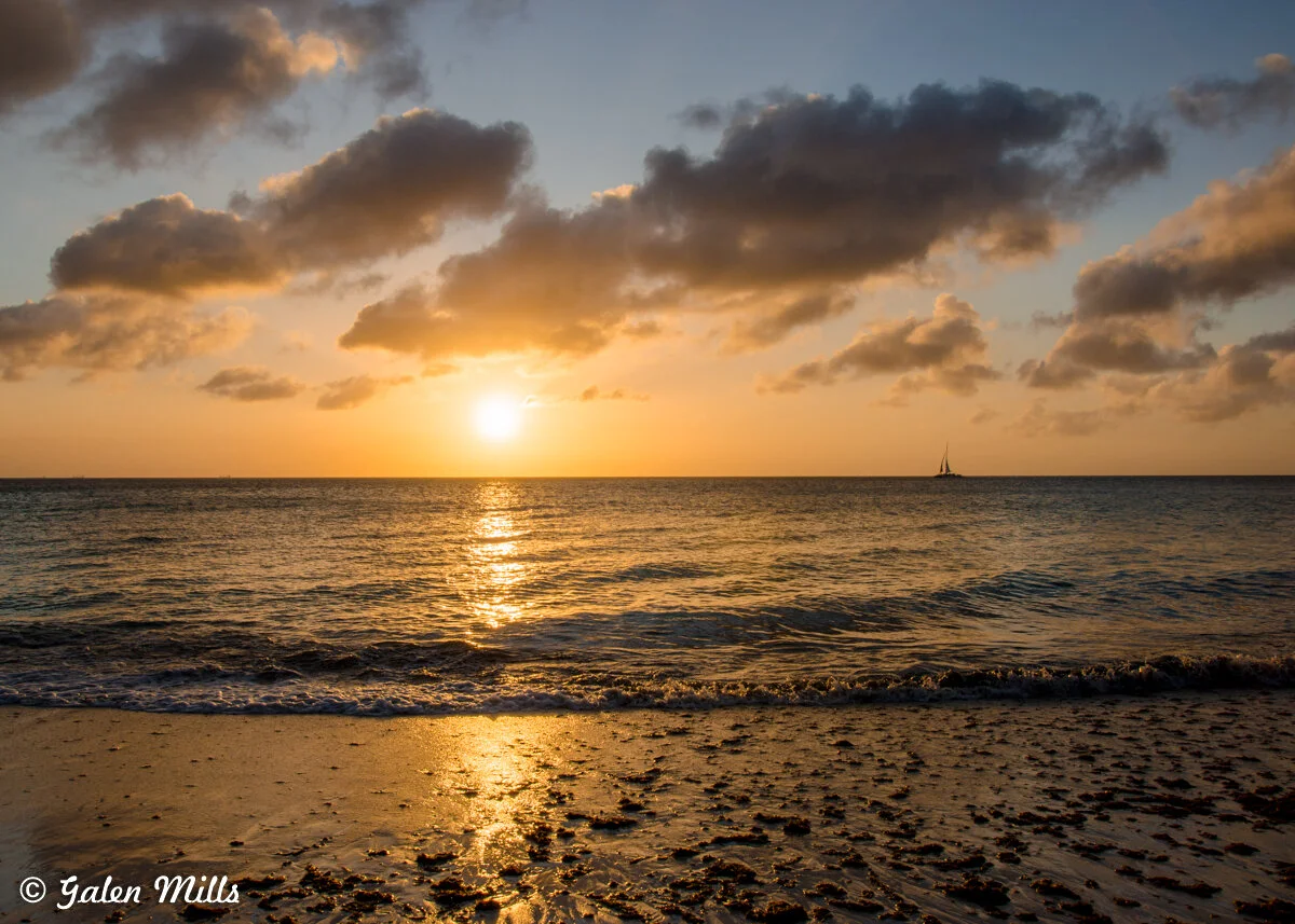 Sunset over ocean with scattered clouds and sailboat silhouette on the horizon.