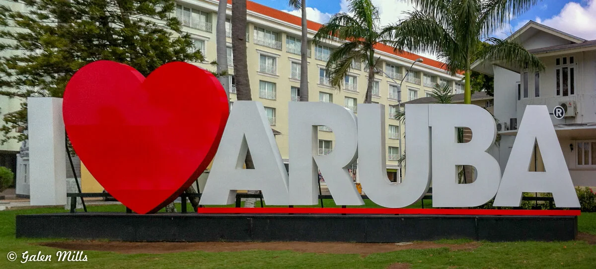 "I Love Aruba" sign with large red heart and white letters in an outdoor setting, surrounded by palm trees and a building in the background.