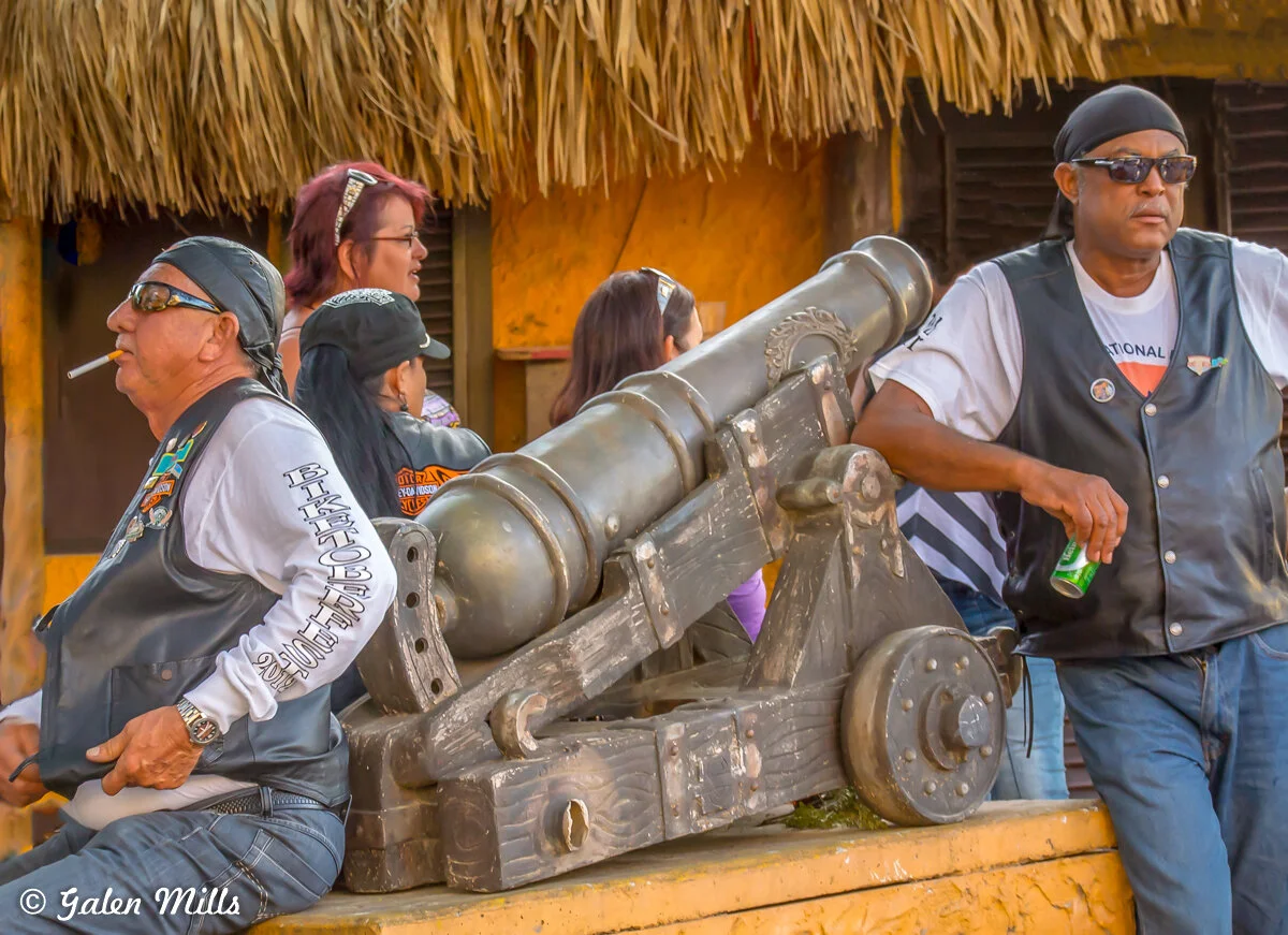 A group of people in a tropical setting, with two men wearing black vests and sunglasses near a large decorative cannon. The background features a thatched roof and yellow walls.
