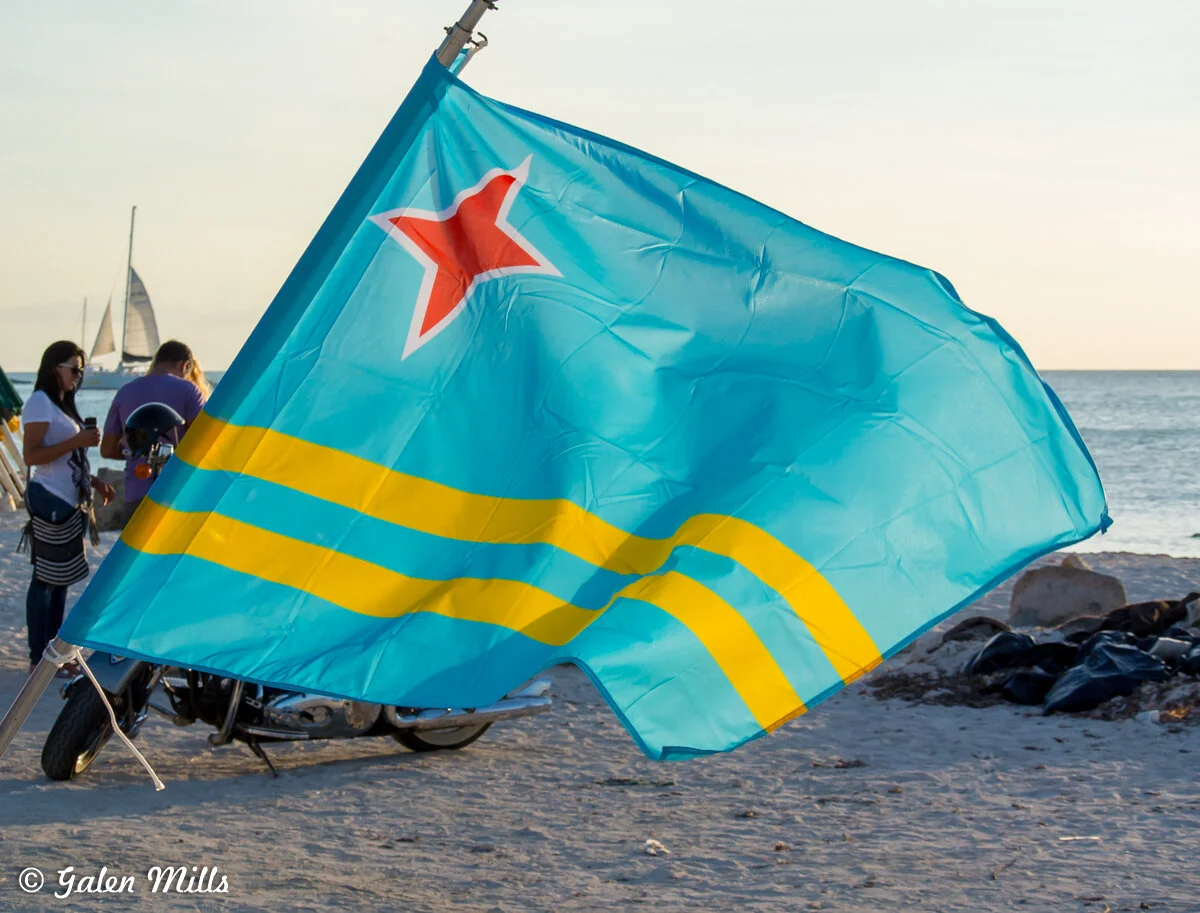 Aruba flag waving on a beach with people and a sailboat in the background.