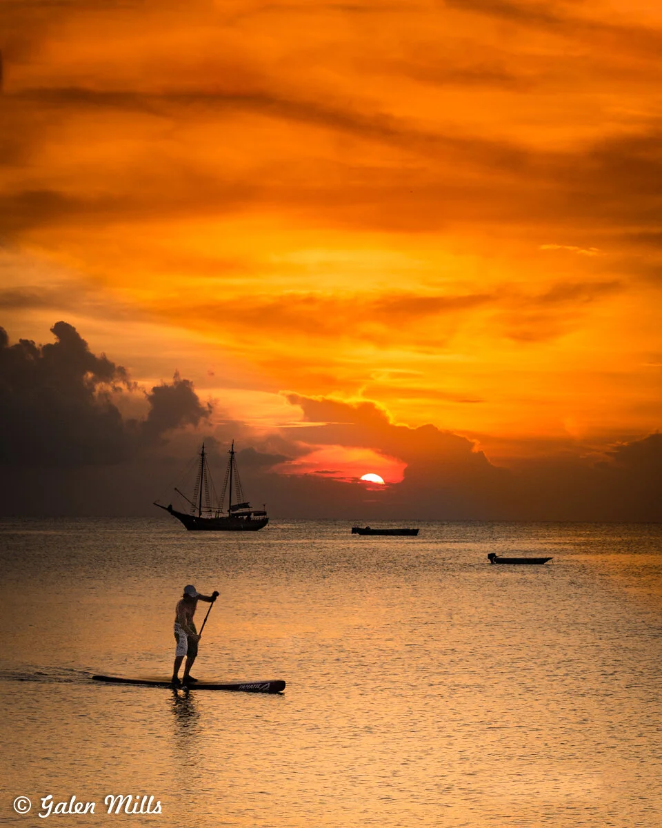 A person paddleboarding on calm ocean water at sunset, with a sailboat and two smaller boats in the background, under a vibrant orange sky.