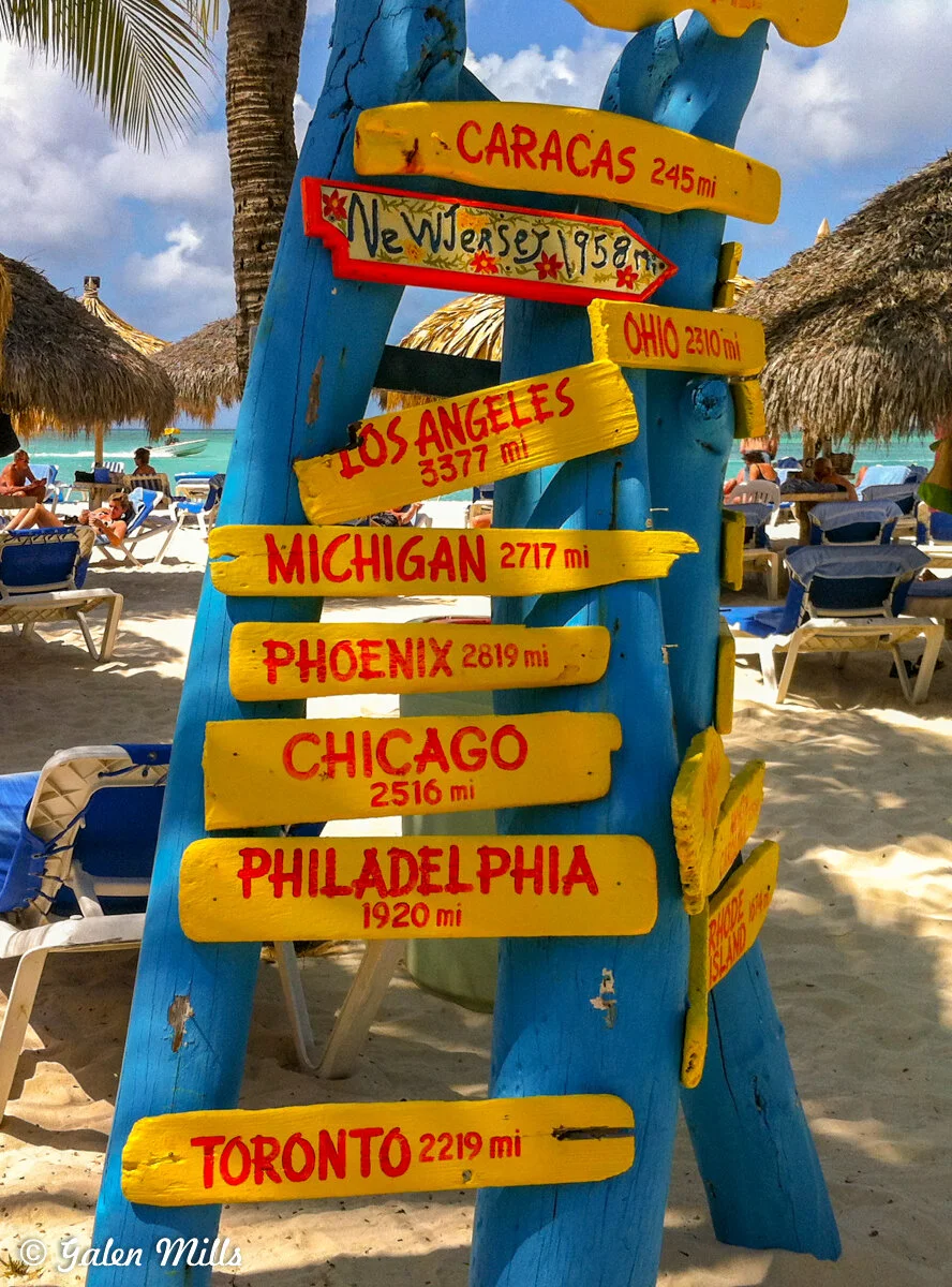 Beach scene with colorful directional signpost, pointing to various cities with distances in miles, and thatched umbrellas in the background.