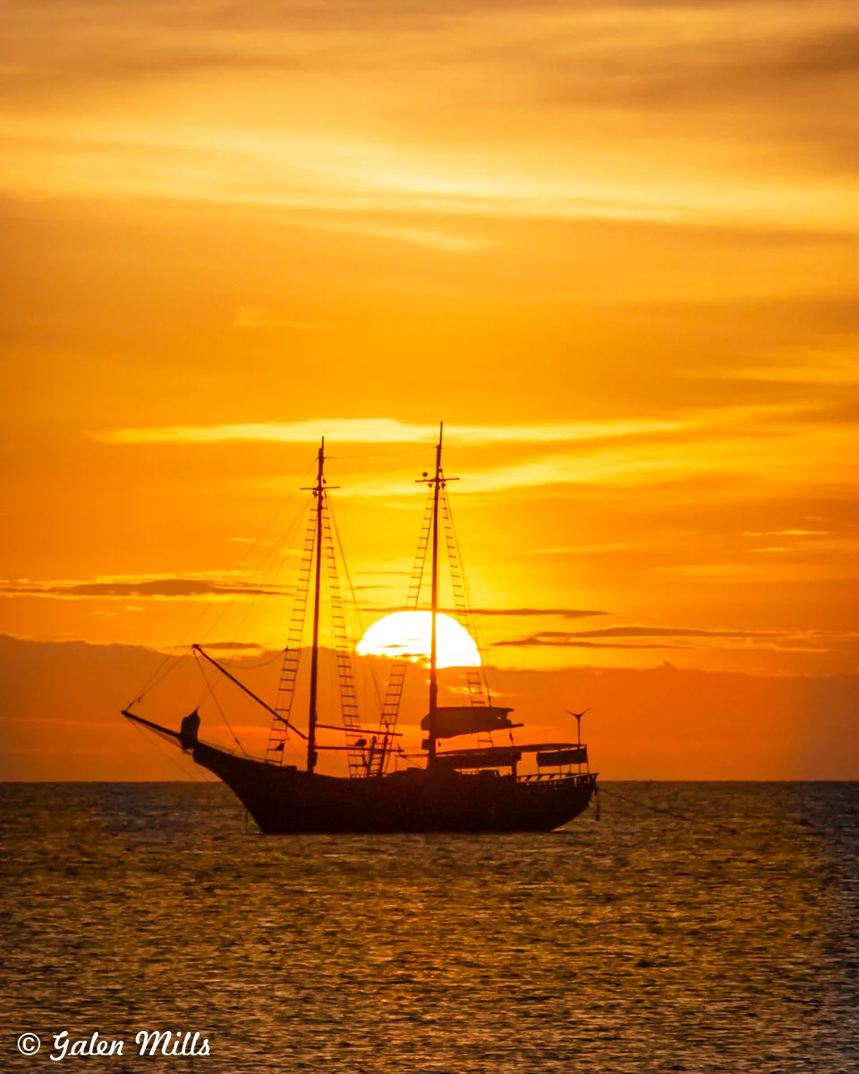 Silhouette of a tall ship at sea during sunset with bright orange sky.