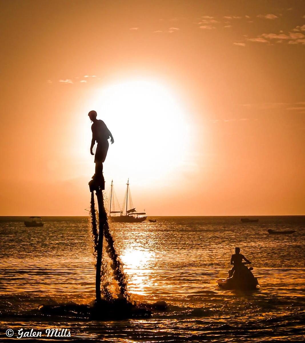 Silhouette of a person flyboarding over the ocean at sunset with a jet ski nearby and a sailing ship in the background.
